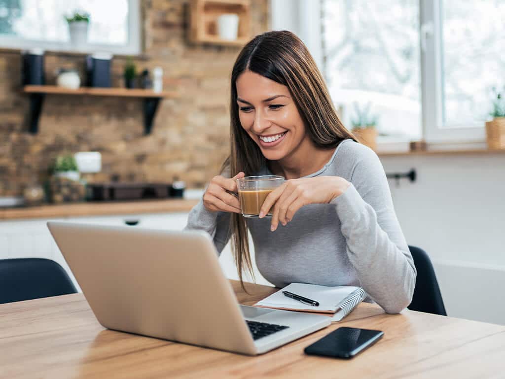 Kobieta pracująca zdalnie, pijąca kawę w kuchni przed laptopem