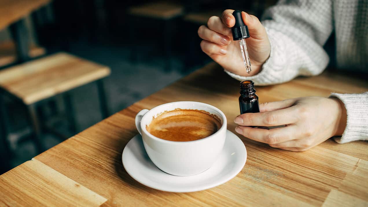 Na stoliku w ciepłej tonacji drewna, stoi biała filiżanka z czarną kawą, a nad nią damskie dłonie wkraplają pipetą olejek CBD z ciemnej buteleczki